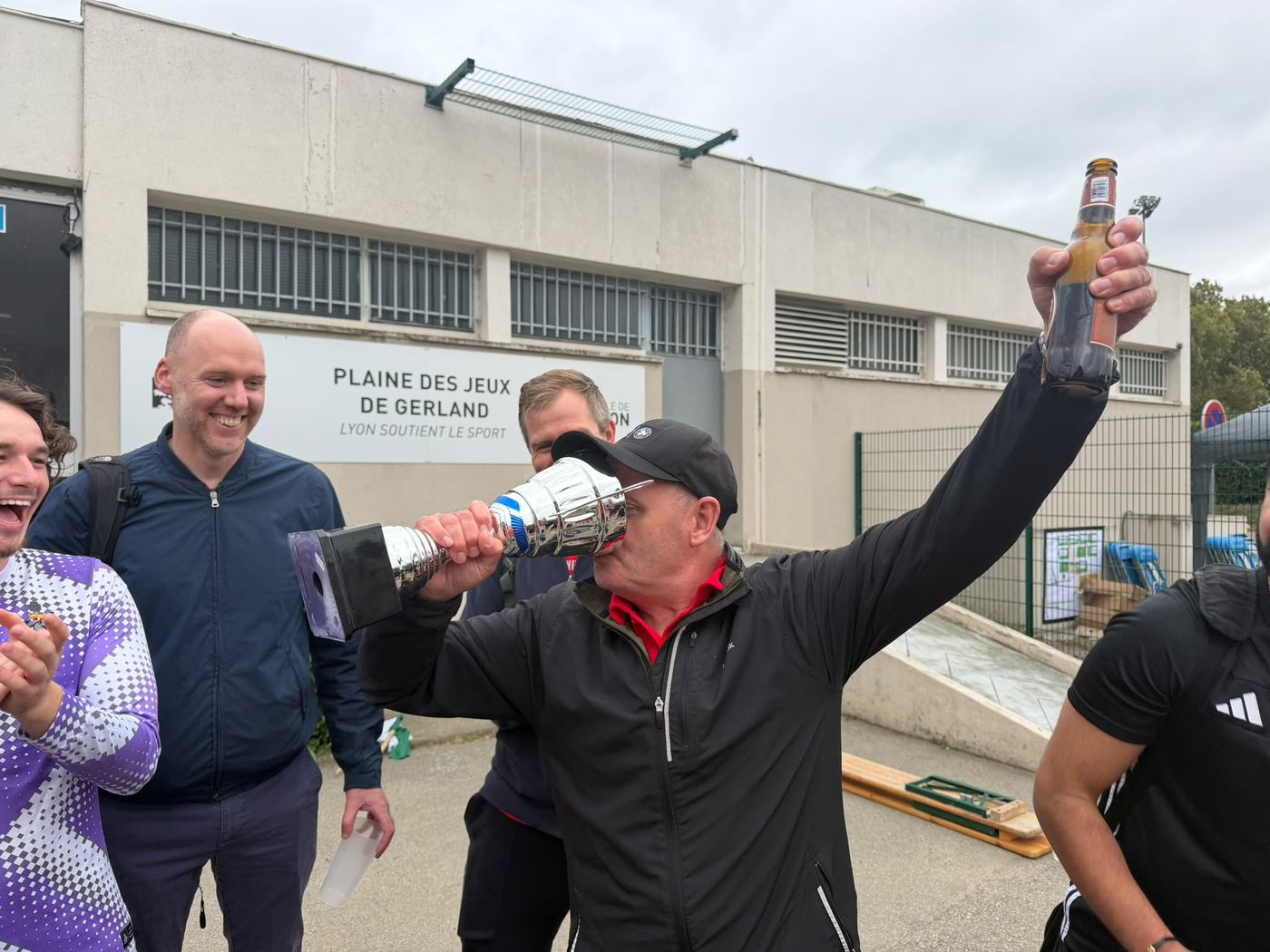 Andy drinking from the silver cup outside the Plaine des Jeux de Gerland in Lyon, bottle raised, as teammates laugh and cheer