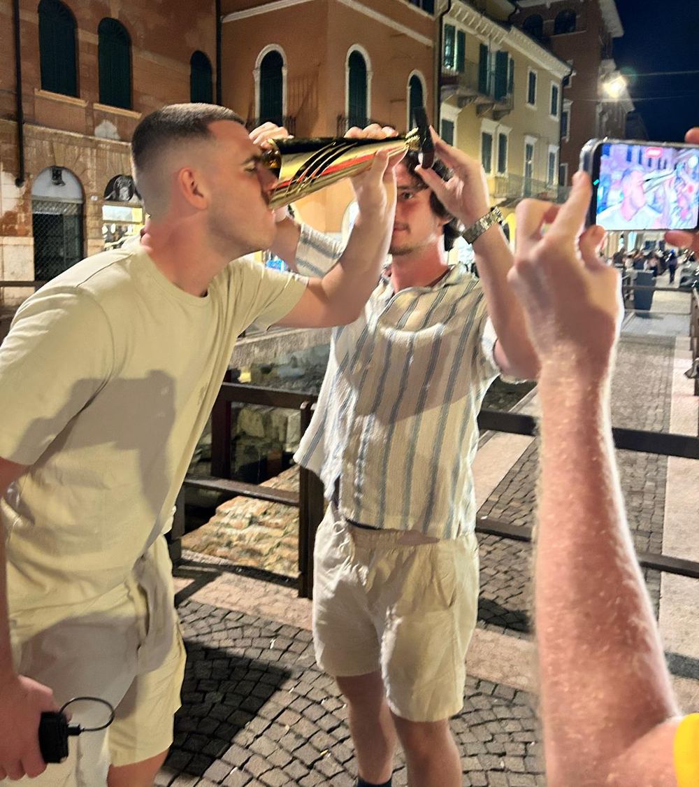 A player drinking from the trophy while a teammate tips it and others film on their phones